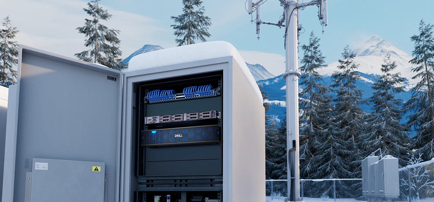 An outdoor telecom enclosure housing rack equipment, with a cell tower and snow-covered mountains behind