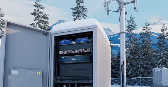 An outdoor telecom enclosure housing rack equipment, with a cell tower and snow-covered mountains behind