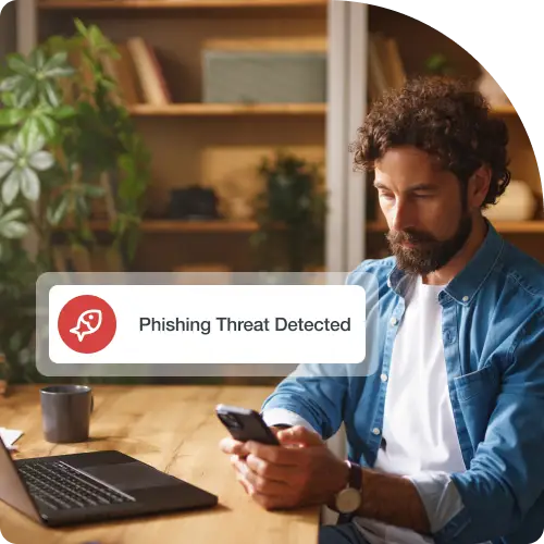 A man working on a laptop at his home office is looking at his mobile phone. A digital message box displays a Phishing icon and the text Phishing Threat Detected.