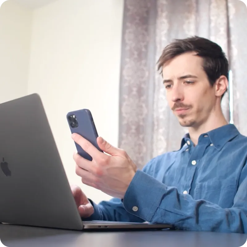 Person in blue shirt at desk using laptop and holding smartphone