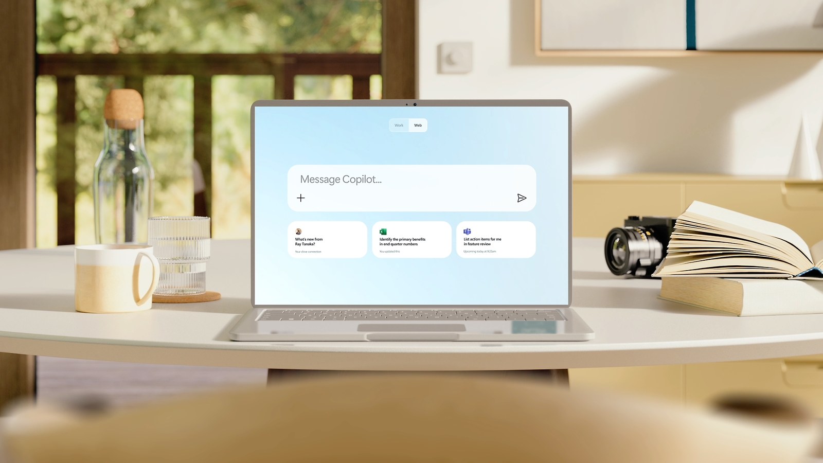A laptop on a desk showing Copilot Chat with a &ldquo;Message Copilot&hellip;&rdquo; box, Work and Web tabs, and three suggestion cards below it.