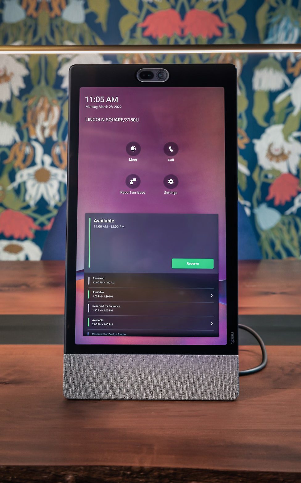 A Microsoft Teams display device standing on a desk showing a call screen, in front of a patterned wall.
