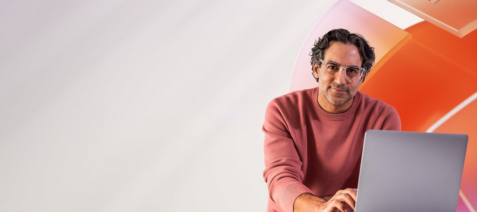 A person in a dusty-pink sweater and clear-framed glasses works at a laptop, with curved orange and pink shapes behind them.