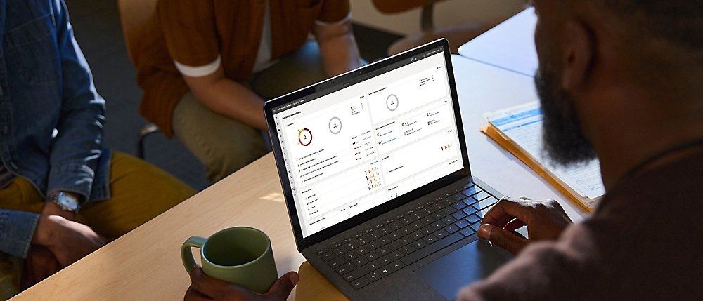 A person with a green mug uses a laptop displaying data dashboards, while two others sit nearby collaborating.