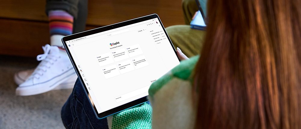 A person in a green sweater holds a tablet displaying a Copilot app.