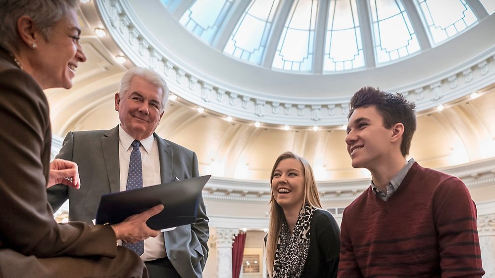 Four people talking together beneath the glass dome of a government building rotunda, one of them holding a folder.