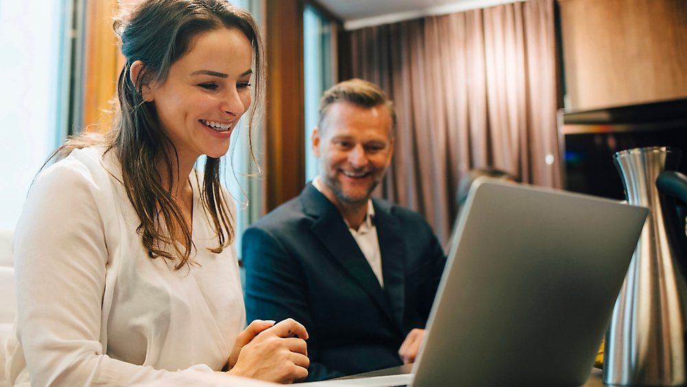 Two people smiling at a laptop screen together in a wood-panelled office.