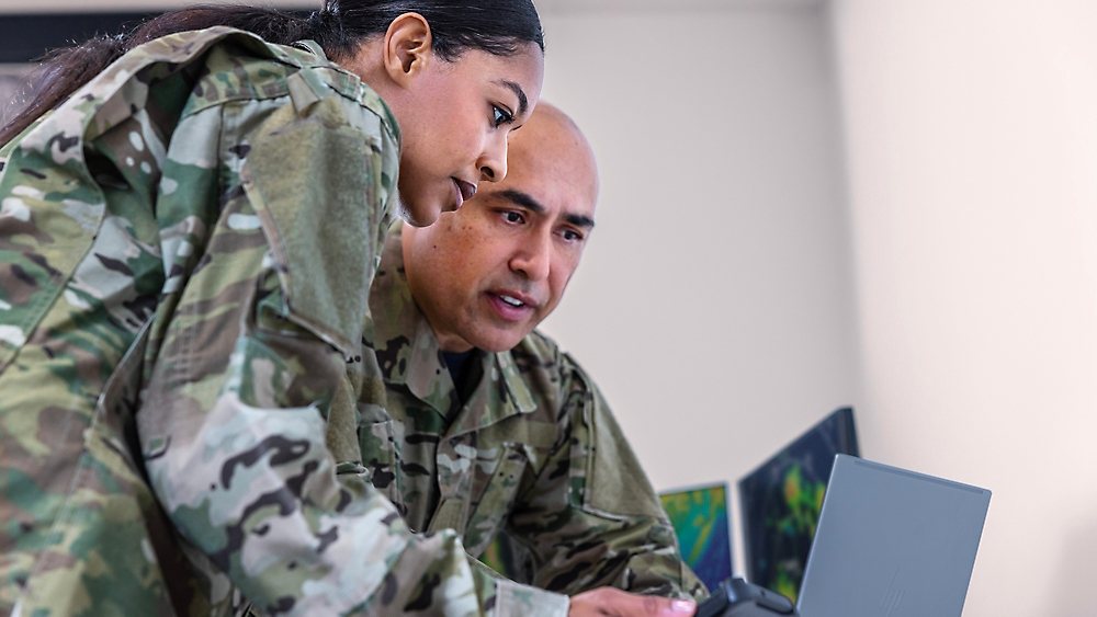 Three service members in camouflage uniform looking at a laptop screen together.